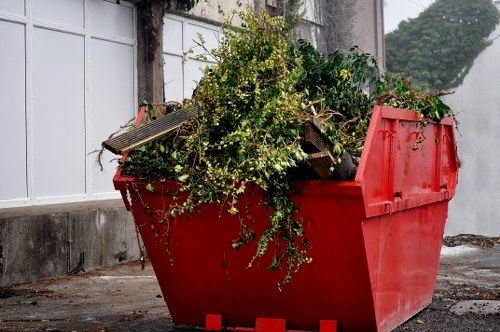 A red skip filled with garden waste, including leafy branches, small twigs, and some wooden planks, positioned on a paved outdoor surface near a grey concrete wall and white fencing. The garden waste appears freshly collected, with green leaves and brown stems spilling over the top of the skip. The scene is set in an area that suggests a backyard or outdoor space in Teddington, with diffused natural lighting indicating an overcast day. The skip's bright red colour contrasts with the greenery of the waste and the muted tones of the surrounding environment. Office Clearance Teddington services may handle such garden waste removal for local clients, with the skip situated for efficient loading or disposal. The image emphasizes the task of clearing and managing outdoor rubbish, consistent with professional rubbish removal activities in the Teddington area, postcodes TW11 or TW12, serving local residential or commercial properties.