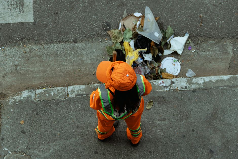 A waste collection vehicle with a predominantly white exterior and red accents is shown from the rear, positioned on a paved street during daylight hours, with sunlight filtering through the trees. Th