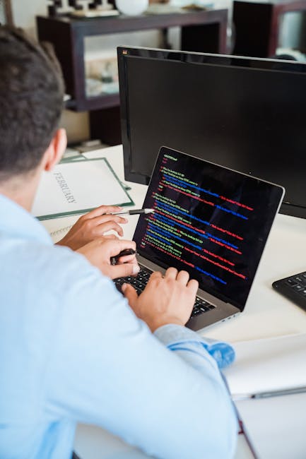 A black laptop with a red-backlit keyboard displaying colorful lines of code on its screen, positioned on a dark surface. To the right of the laptop, there is a white Puma cap with the black Puma logo