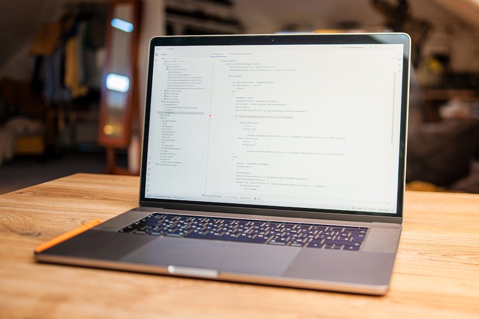 A close-up view of a person's hands using a laptop computer placed on a dark, reflective surface. The laptop screen displays a dark-themed coding or software development interface, with lines of multi