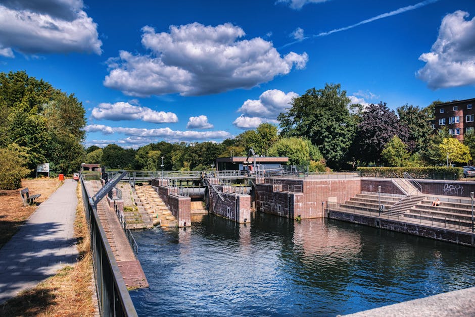 The image depicts a narrow, paved pathway running alongside a canal with several narrowboats moored along the water's edge, some featuring solar panels on their roofs and small outboard motors. In the