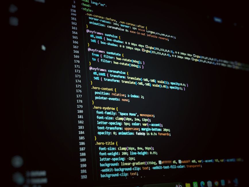 A close-up view of a computer screen displaying lines of multicoloured programming code on a dark background, with text in red, green, blue, and purple. The code appears to be HTML or CSS, featuring t